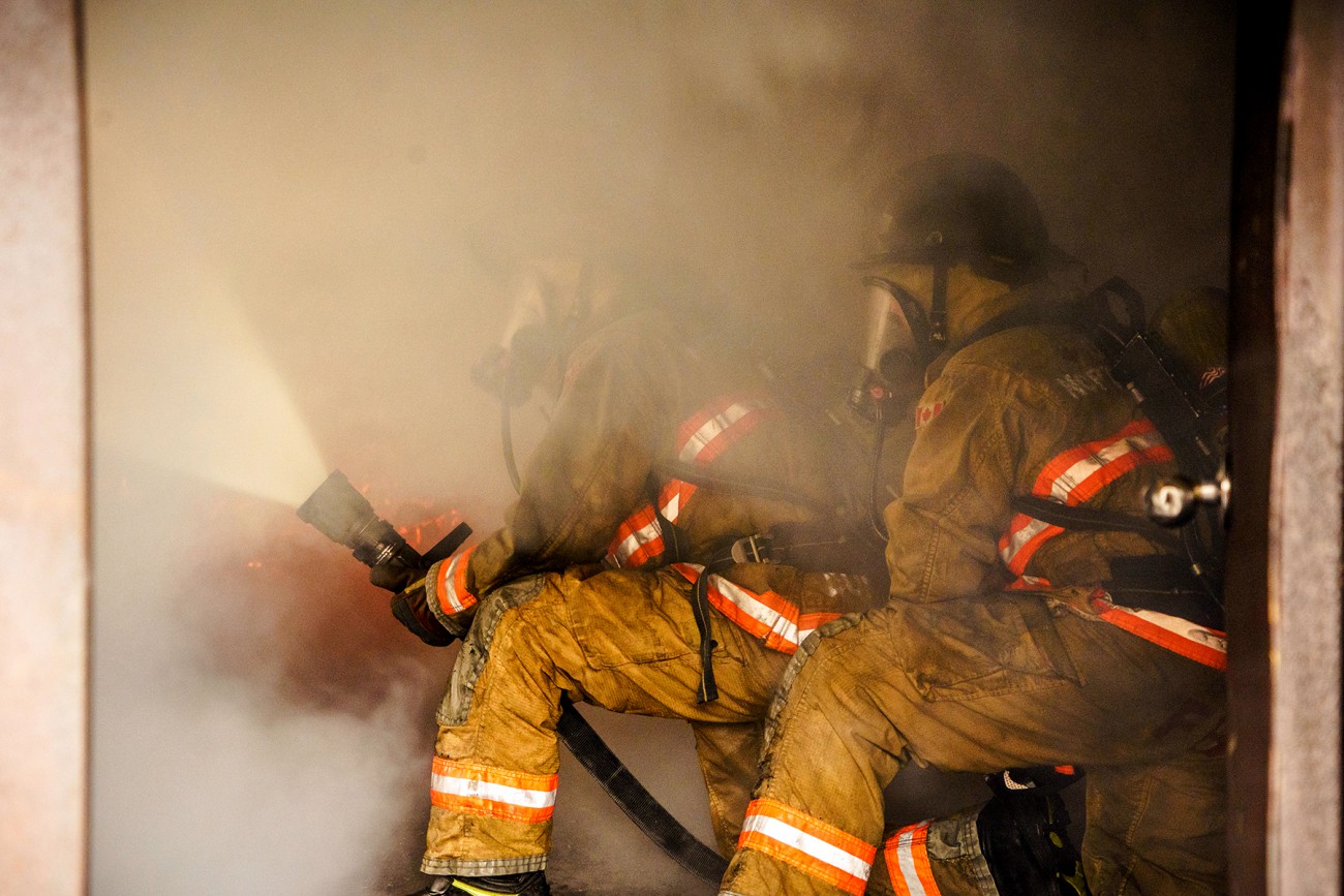 Two firefighters in a smoky room using a hose to battle a fire. 