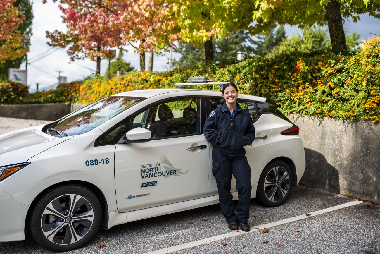 A smiling female bylaw officer stands in front of a white electric fleet car.