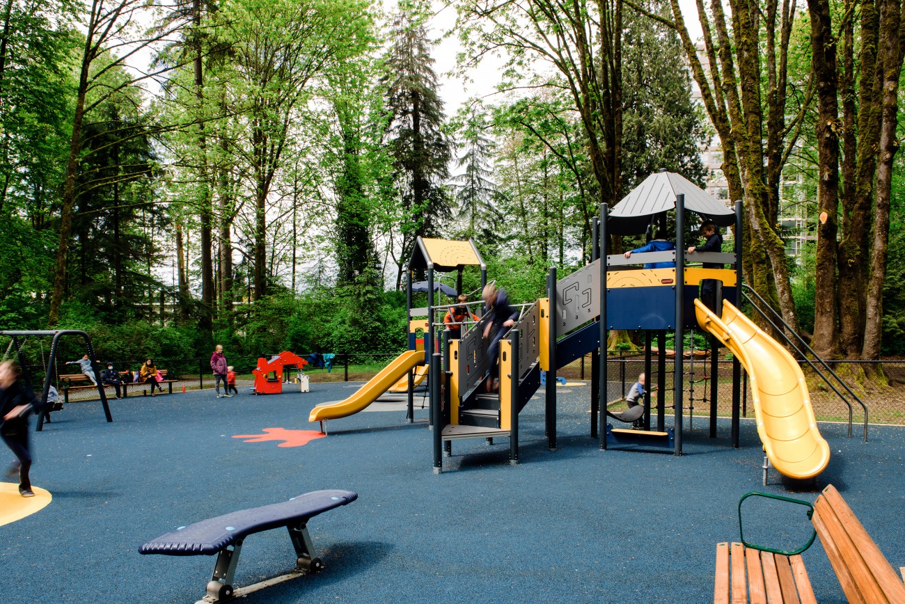 Kids of different ages play at a colourful playground with slides and swings and other features. The playground is surrounded by tall trees.