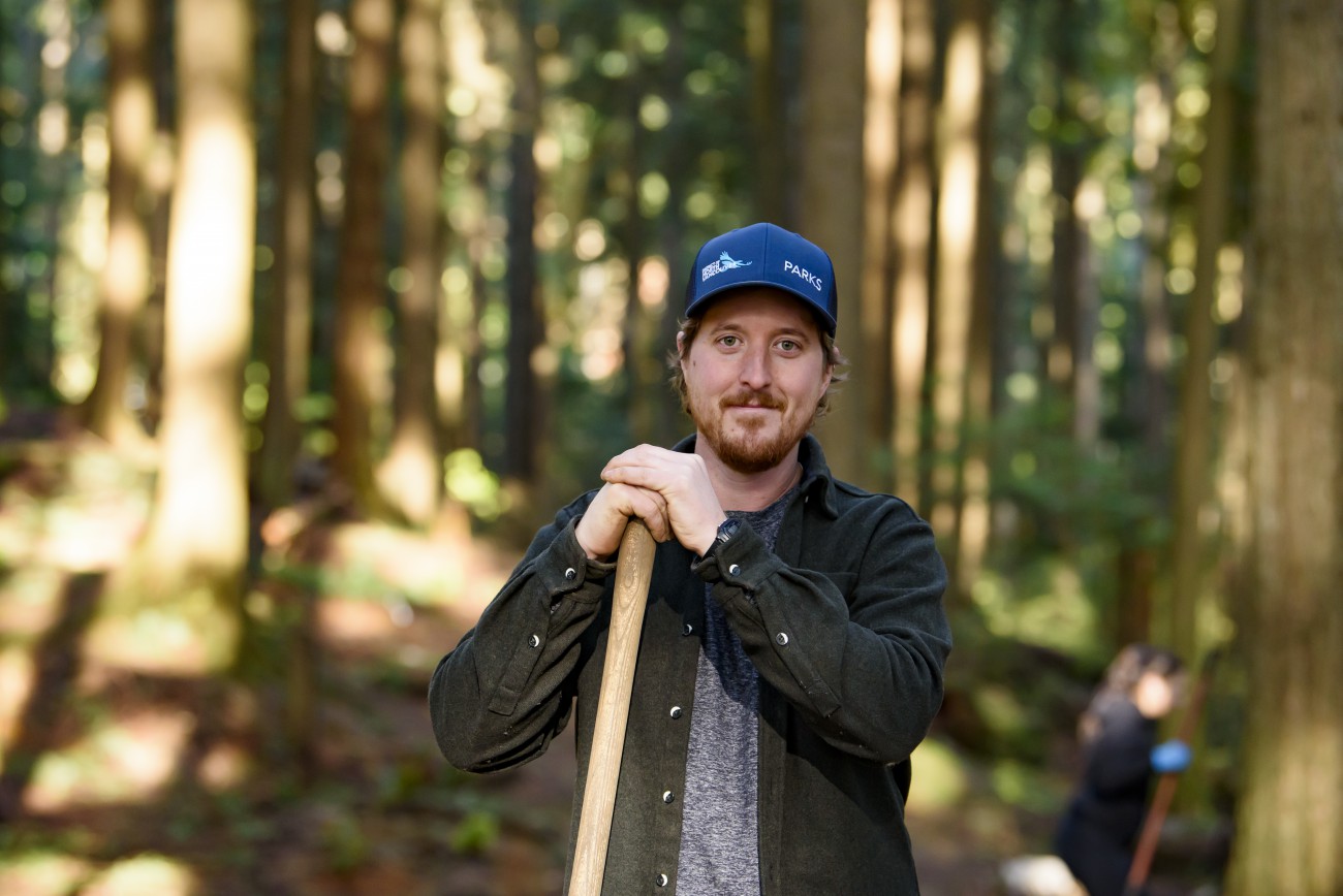 A smiling bearded man wearing a blue cap with the word Parks holds a shovel against a blurred forest background.