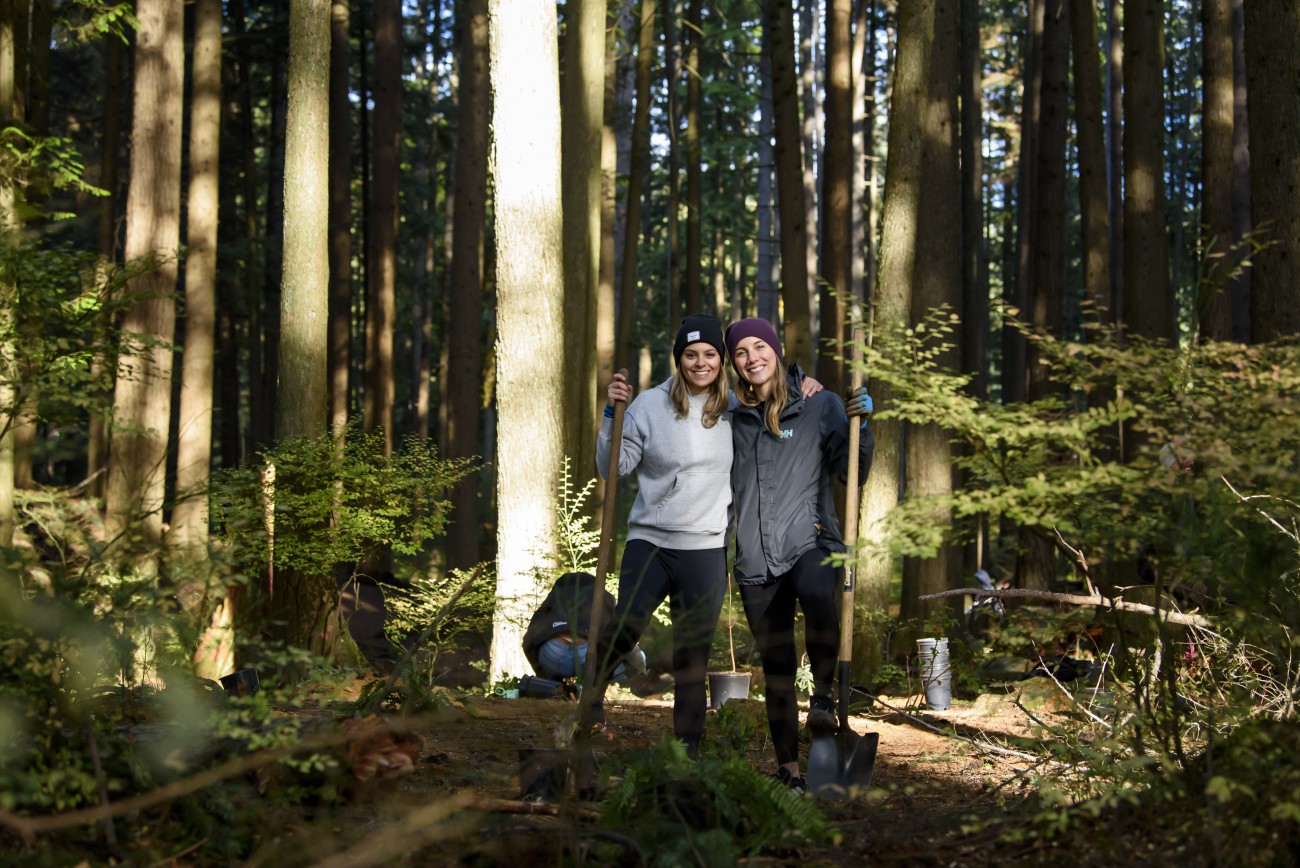 Two smiling women in toques holding shovels participate in a shrub planting event. They stand against a forested background. 