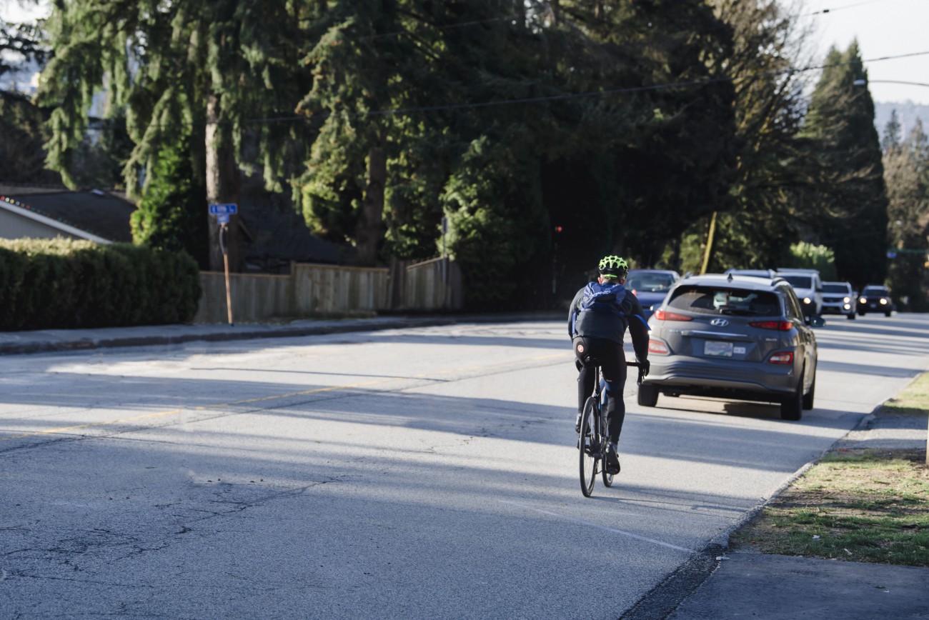 A cyclist rides down a tree-lined street during a morning commute as vehicles drive along the two-land road. 