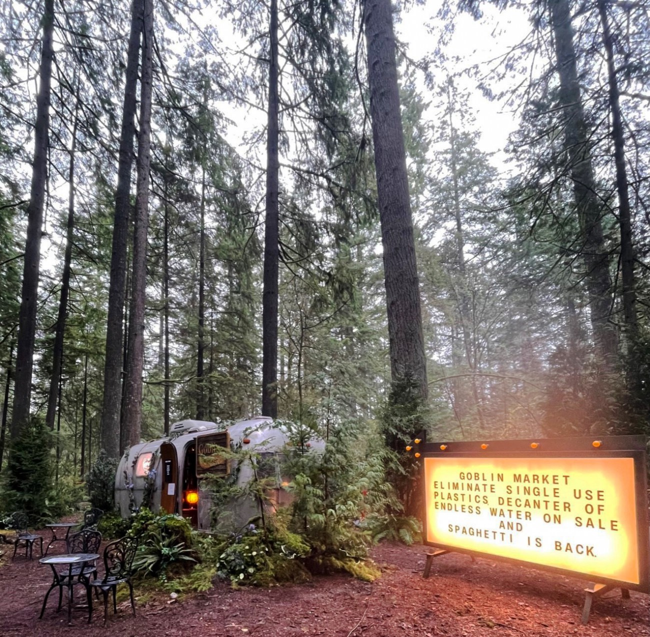 An Airstream trailer is parked in the woods with a chair sitting in front of it. A glowing sign reads: Goblin Market. 