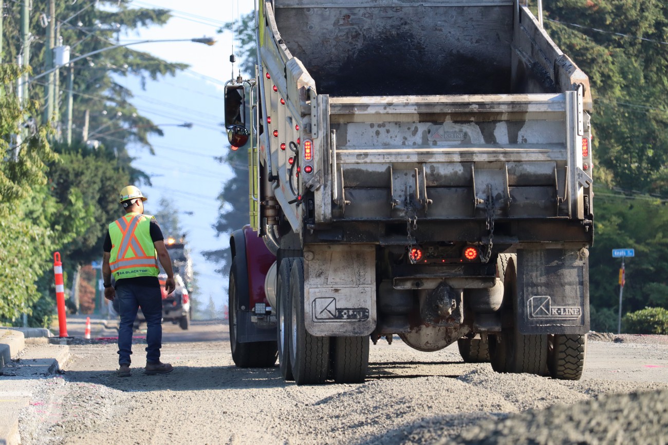 A worker in a yellow hardhat walks beside a dump truck on a road that is being paved.