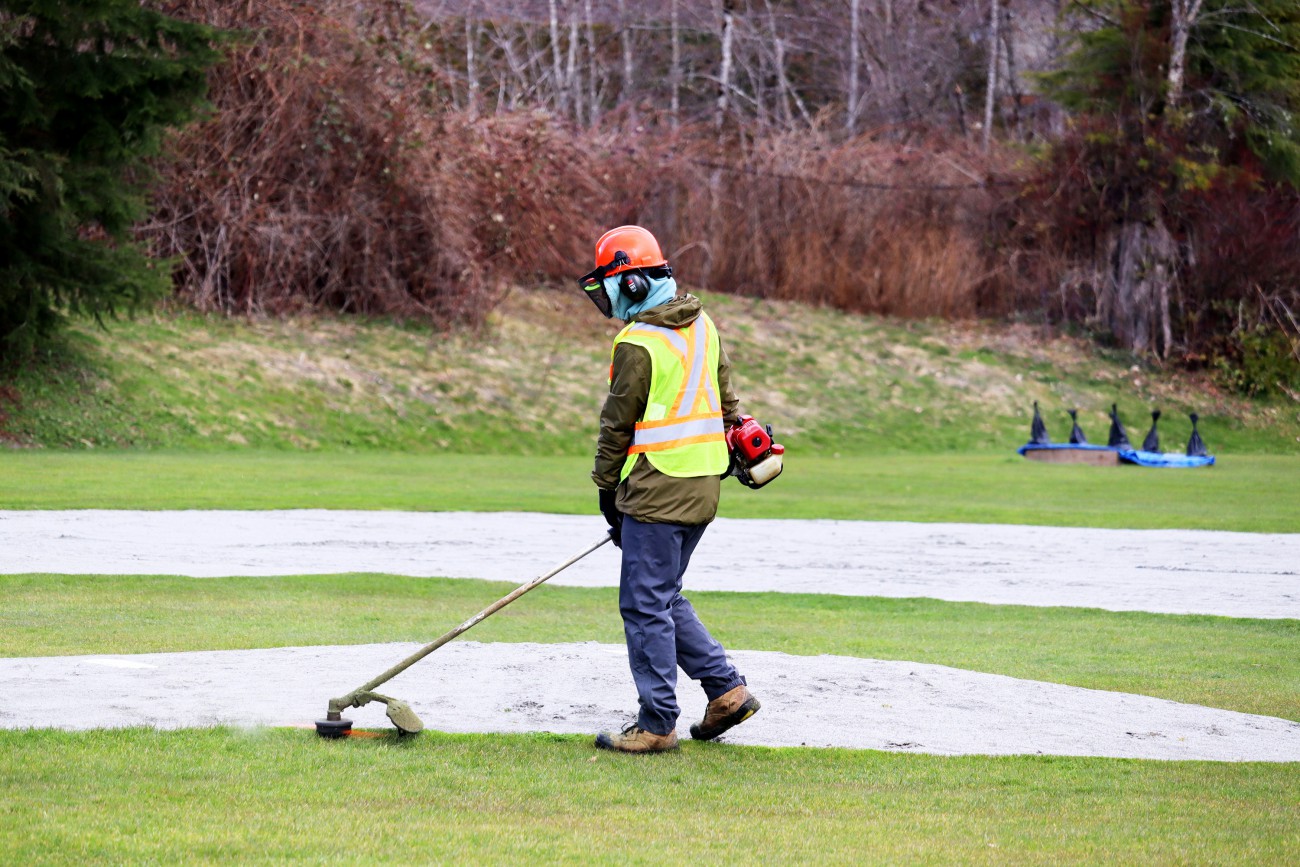 A Parks crew member uses a weedeater to trim the grass around the pitching mound of a baseball field.