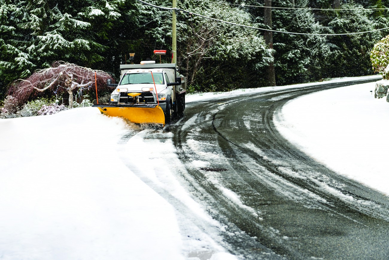 A white snowplow removes snow on a tree-lined street. 