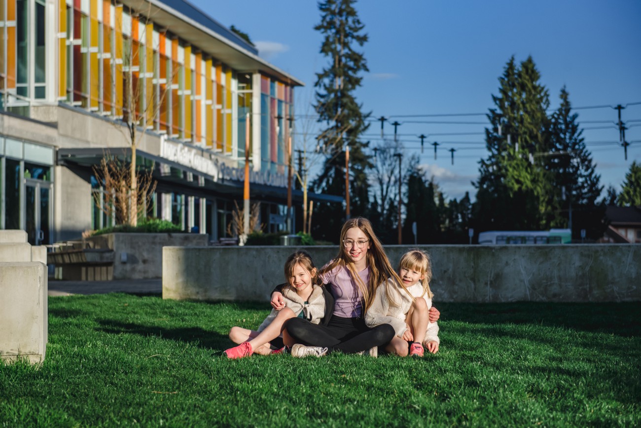 Three sisters sit on the grass together smiling with a new community rec centre in the background.