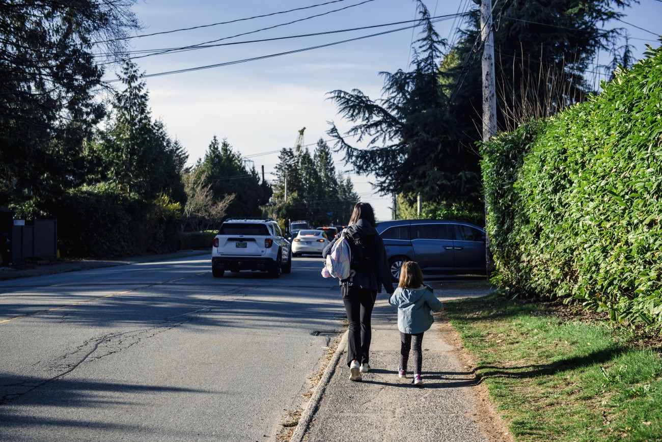 A mother and daughter walk down a sidewalk hand in hand as traffic passes by on a busy street. 