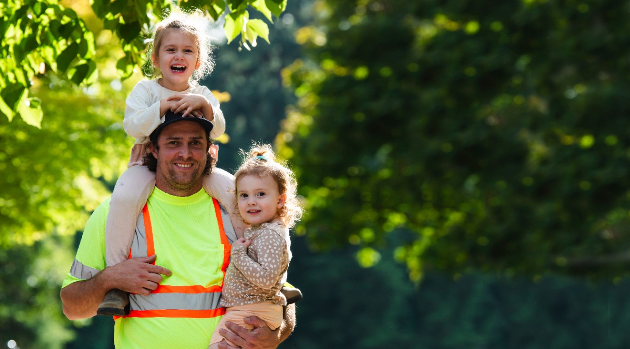 A father wearing a high-visibility shirt holds his two young smiling daughters with a blurred forest backdrop behind them.