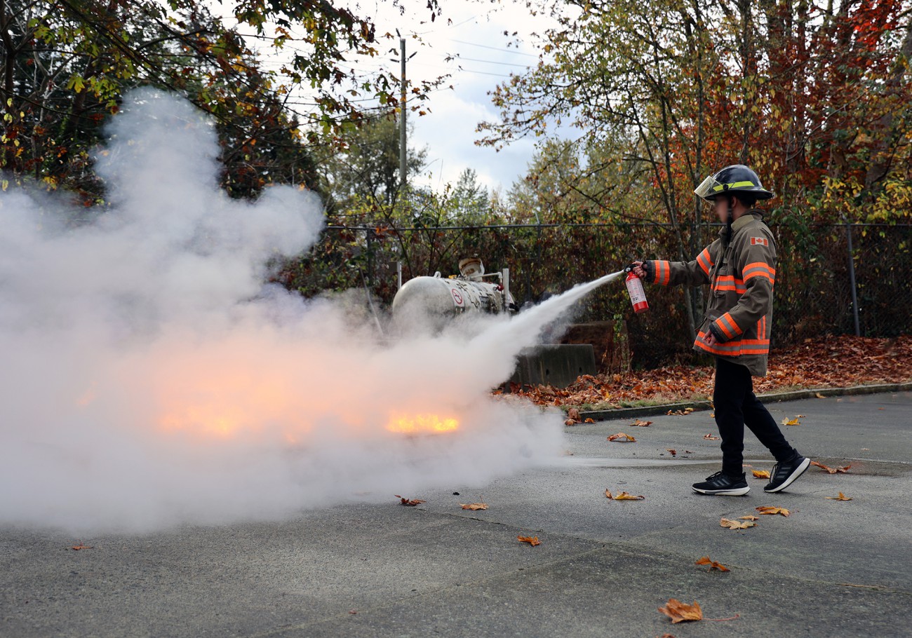 A student wearing firefighter gear uses a fire extinguisher to put out a small blaze.
