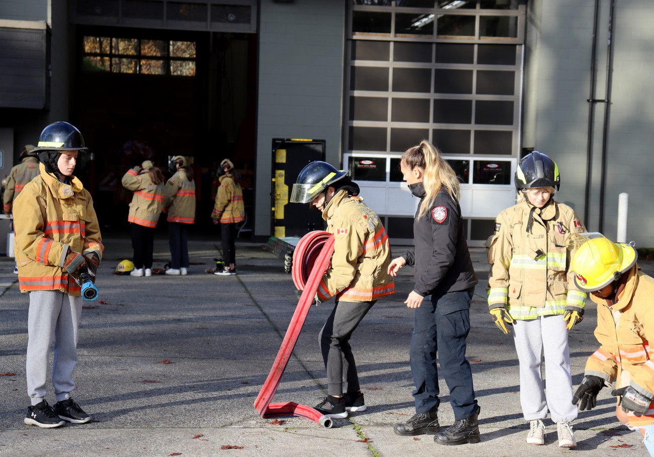 A female firefighter dressed in blue teaches a group of students clad in firefighter gear how to roll up a fire hose.