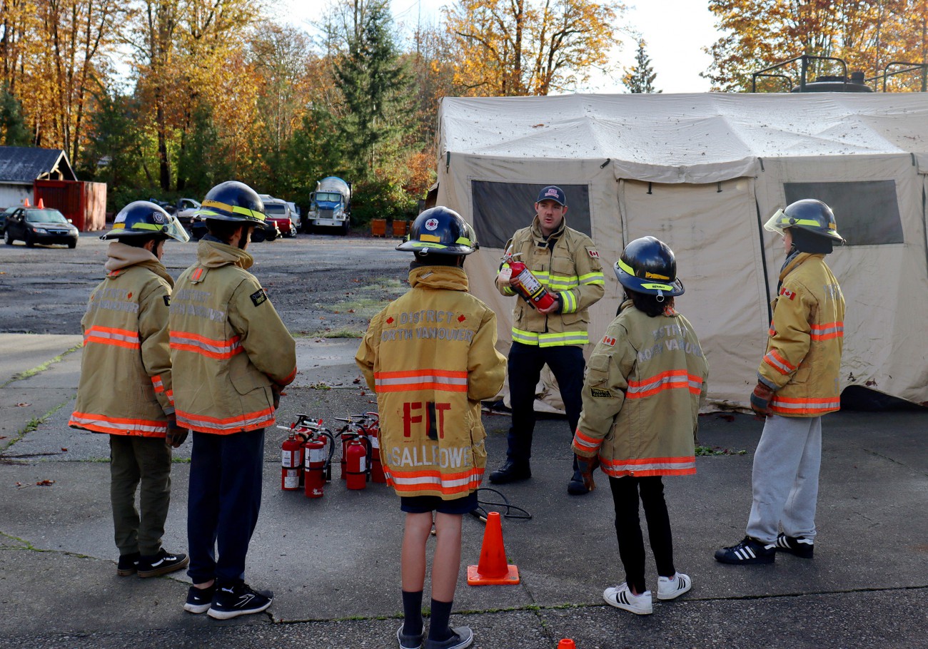 A firefighter in turnout gear demonstrates how to use a fire extinguisher as a group of students wearing firefighter gear watch.