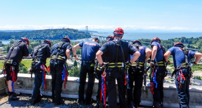 A group of firefighters clad in blue uniforms look over the edge of a tall building.