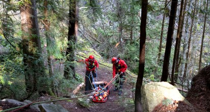Two red-uniformed firefighters practise transporting an injured hiker in the woods.