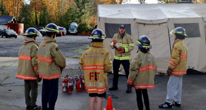A firefighter in turnout gear demonstrates how to use a fire extinguisher as a group of students wearing firefighter gear watch.