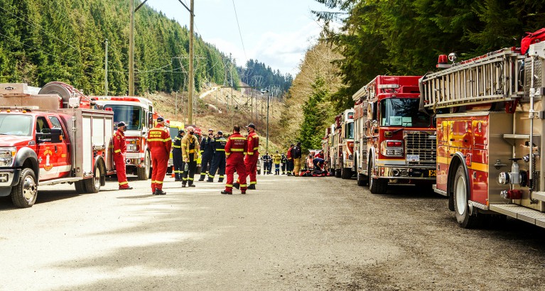 A long line of fire response vehicles parked on a gravel street as a group of firefighters discuss training plans.