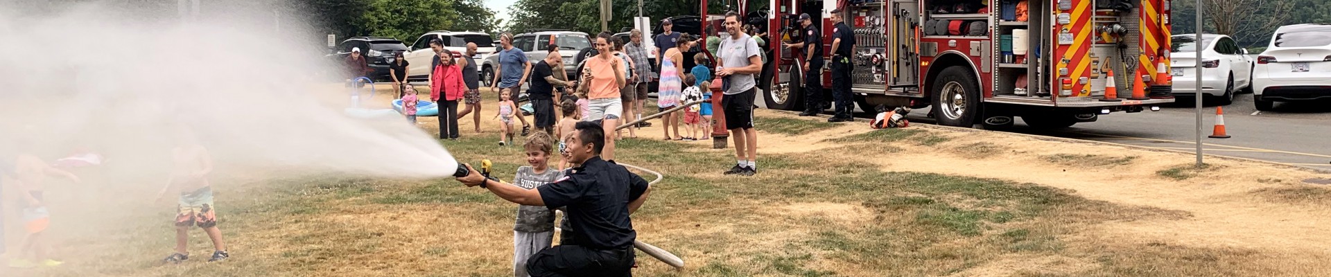 A smiling firefighter stands beside a young boy who is practising using a firehouse in the park. Other community residents are smiling and talking with firefighters in the background.