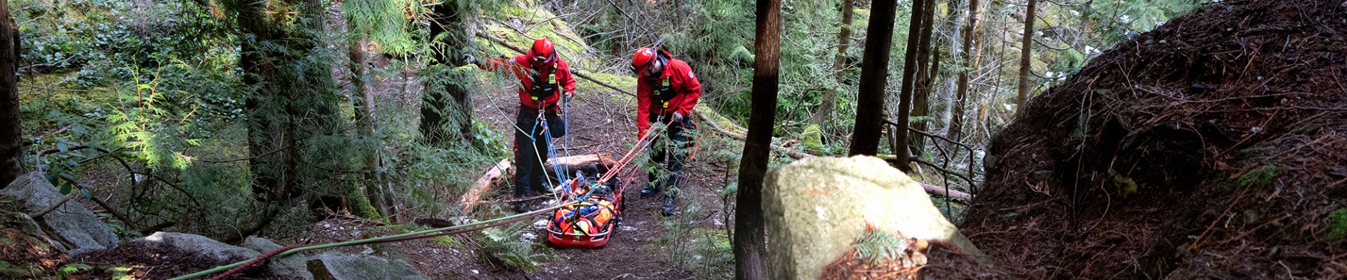 Two red-uniformed firefighters practise transporting an injured hiker in the woods.