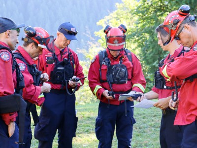 A group of DNVFRS firefighters in wildfire fighting gear stand in a circle coordinating radios