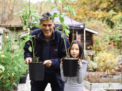 A father and daughter with smiles on their faces hold up potted trees in an outdoor nursery.
