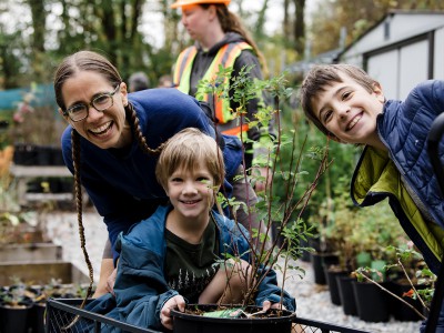 A smiling woman and two young pick up a potted shrub from a nursery as part of the District of North Vancouver's urban tree program.
