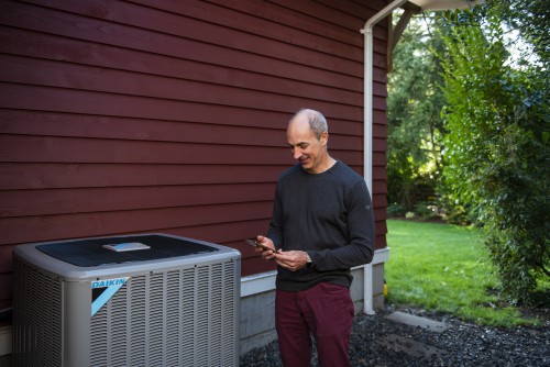 A man stands outside his home in front of a heat pump.