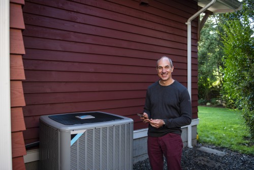 A smiling man stands beside a heat pump outside his home.