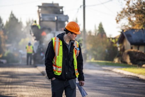 A man in an orange hard hat inspects a freshly paved street.
