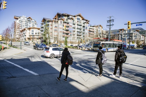 Three young people with backpacks stand at a crosswalk waiting for the light. In the intersection, cars and a bus move by. In the background, a cluster of new low-rise condo buildings.